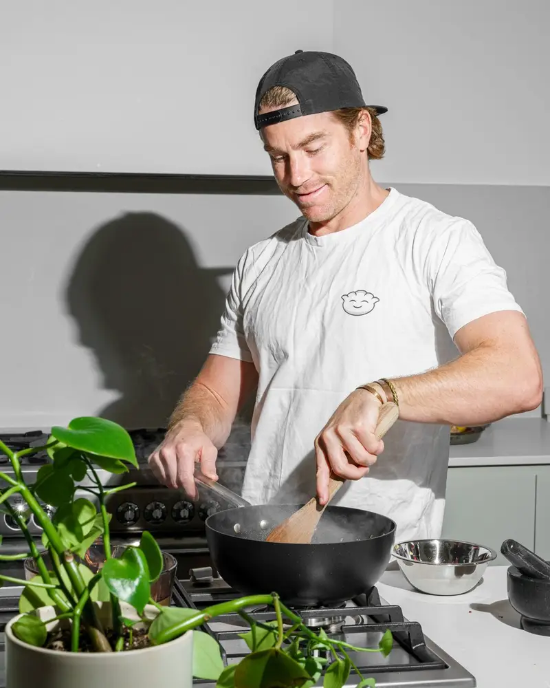 Hayden smiling as he stirs a black wok with a wooden spatula, a stainless bowl and stone mortar beside him