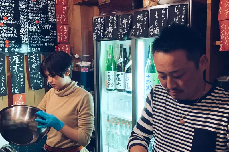 Behind the counter at Sakaba Ikura Mokuzai in Kyoto, with chalkboard menus and the sake fridge